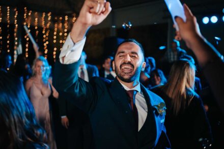 El novio grita con el puño en alto en la pista de baile, con una cortina de luces detrás