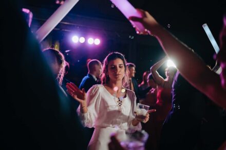 La novia baila con los ojos entornados y un vaso en la mano entre invitados con barras de luz