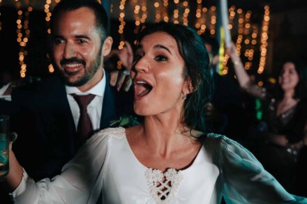 La novia canta con la boca muy abierta y el novio sonríe detrás de ella, con luces al fondo