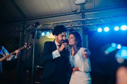 La novia sonríe abrazada a un hombre de pelo rizado que canta al micrófono en el escenario