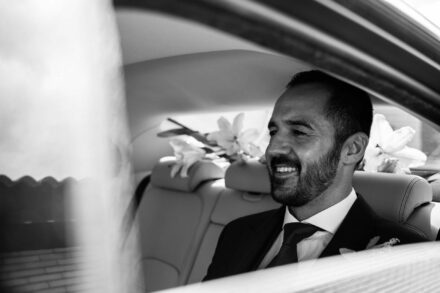El novio sonríe en el asiento trasero del coche, con flores blancas detrás, en blanco y negro