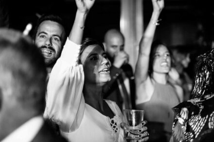 La novia canta con un brazo en alto y un vaso en la mano junto al novio, en blanco y negro