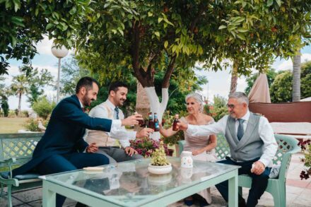 El novio y tres invitados brindan con botellines de cerveza alrededor de una mesa de cristal bajo un naranjo