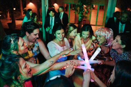 La novia y varias invitadas, una con peluca rosa, juntan manos y barras de luz en el centro del corro