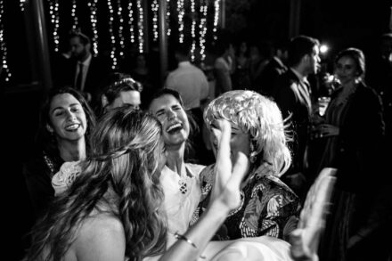 La novia ríe a carcajadas abrazada por invitadas, una con peluca rubia, en blanco y negro