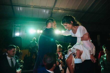 A hombros de los invitados, el novio sujeta del brazo a la novia, que le sonríe, con el grupo al fondo