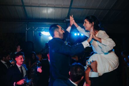 Los novios, cada uno a hombros de los invitados, chocan las manos en alto en la pista de baile