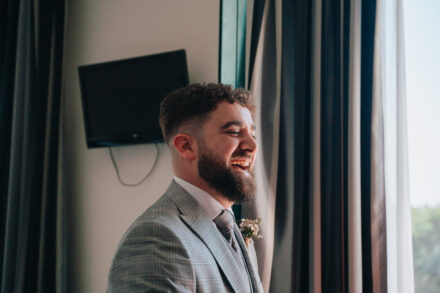 The groom laughs by the bedroom window, backlit, with the buttonhole already on his lapel
