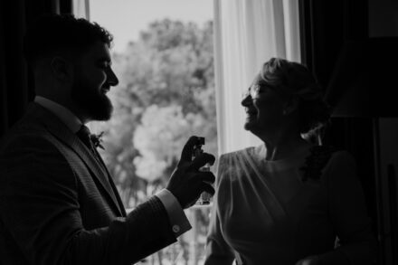 The groom puts on cologne facing a woman in glasses, both backlit, in black and white