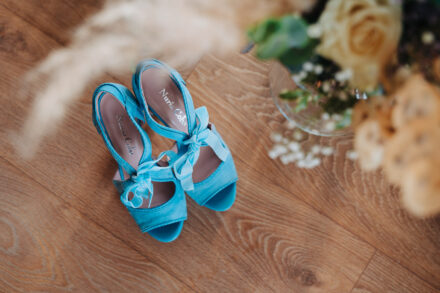 The bride’s blue bow sandals seen from above on the wooden floor beside the bouquet