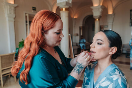 A red-haired make-up artist with a floral tattoo paints the bride’s lips in a lounge with columns