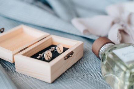 Cufflinks engraved with initials in a small wooden box, beside the groom’s jacket and cologne
