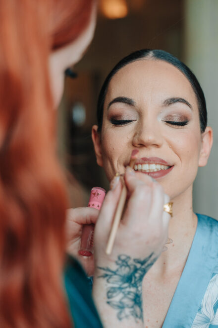 The bride smiles with her eyes closed as a make-up artist outlines her lips
