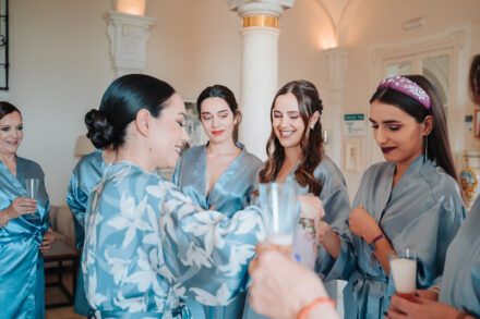 The bride, in a floral robe, hands out glasses to the guests in blue satin robes