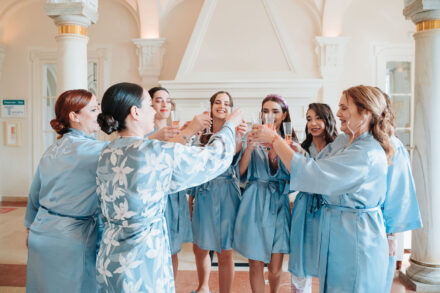 The bride raises a toast with the guests in blue robes in a lounge with columns and a fireplace