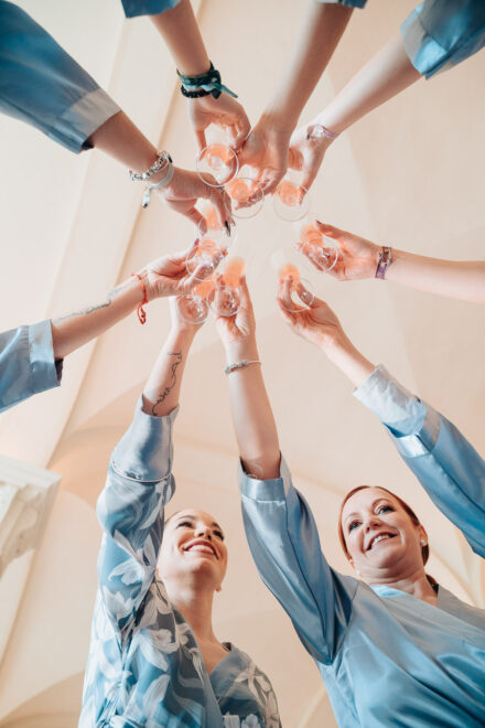 The bride and her guests clink glasses with their arms raised, seen from below