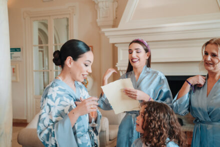 A guest in a headband holds up a letter to the bride, who smiles with a glass in hand