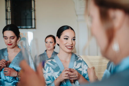 The bride smiles, glass held in both hands, as she listens to the letter, surrounded by guests in robes