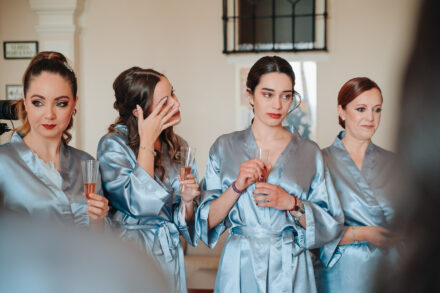 Four guests in blue robes listen, three of them holding glasses; one raises her hand to her eyes