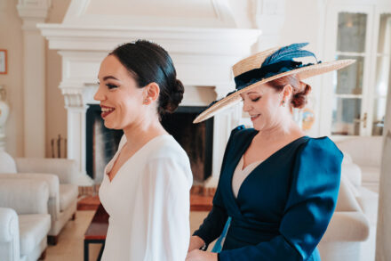 A woman in a straw boater with a blue feather fastens the bride’s dress by the fireplace