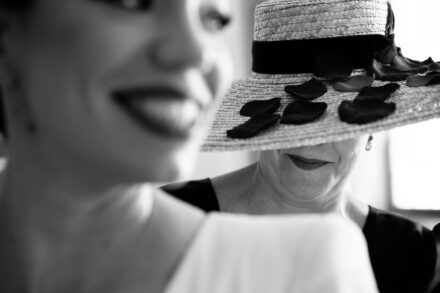 The bride, out of focus, smiles in front of a woman in a petal-trimmed straw hat, in black and white