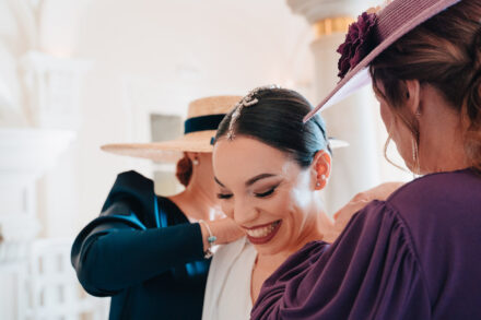 The bride, in a jewelled headpiece, laughs between two women in hats who help her get dressed
