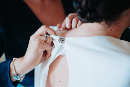 Hands wearing rings fasten the jewelled button at the back of the bride’s dress