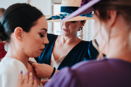 A woman in a straw hat with blue ribbons adjusts the bride’s dress during the preparations at Hotel Legado Oromana