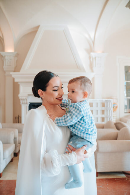 The bride smiles holding a baby in a blue gingham shirt beside a white fireplace