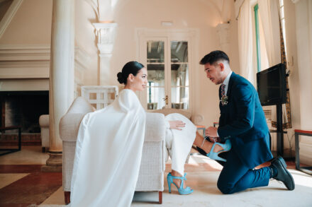 A young man in a navy suit kneels to put blue heels on the bride, seated on an armchair in the lounge