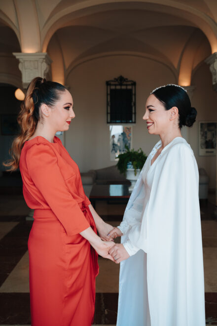 The bride and a guest in a coral-red dress smile at each other, holding hands beneath the hotel’s vaulted ceiling