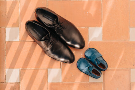The groom’s black shoes next to a pair of tiny blue baby shoes on a tiled floor