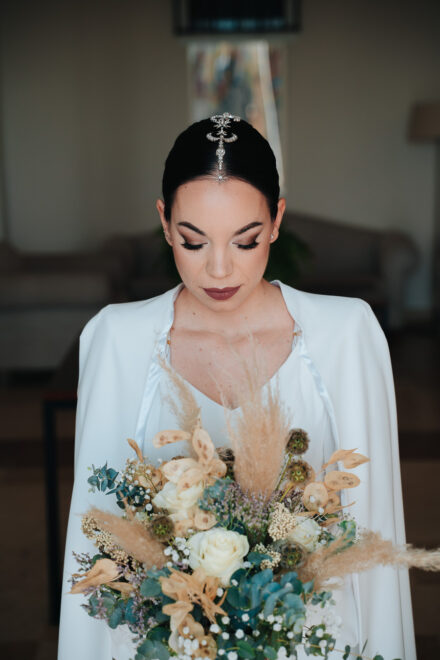 The bride looks down, wearing a jewelled headpiece and holding a bouquet of dried flowers and pampas grass
