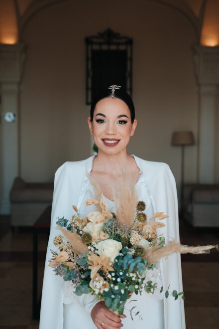 The bride smiles at the camera in her white cape, holding a bouquet of white roses and dried flowers