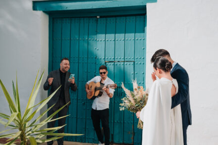 A guitarist plays by a turquoise door as the bride, from behind, raises a hand to her face beside the young man in navy