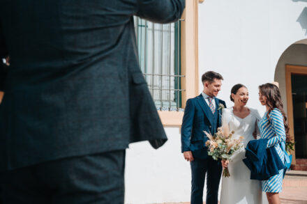 The bride laughs between the young man in the navy suit and a guest in a striped dress, in front of a white facade