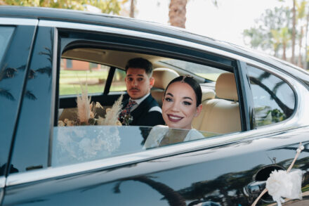 The bride smiles from the back seat of a black car beside the young man in the navy suit, arriving at the ceremony