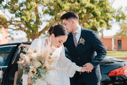 The young man in the navy suit kisses the bride’s head as he helps her out of the car