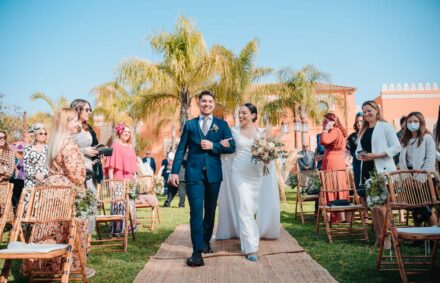 The bride walks down the aisle on the arm of the young man in the navy suit, past standing guests and palm trees