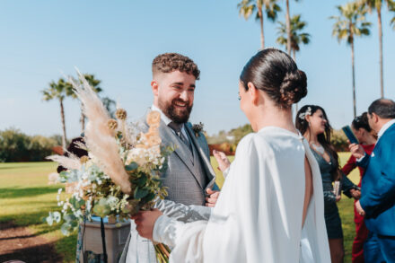 The groom smiles as he welcomes the bride at the altar, seen from behind with her bouquet in hand