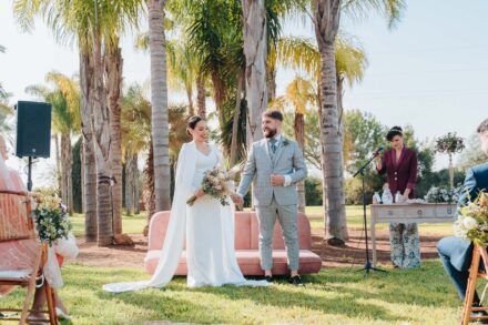 The couple stand before a pink sofa among palm trees, next to the officiant in a burgundy jacket at her table