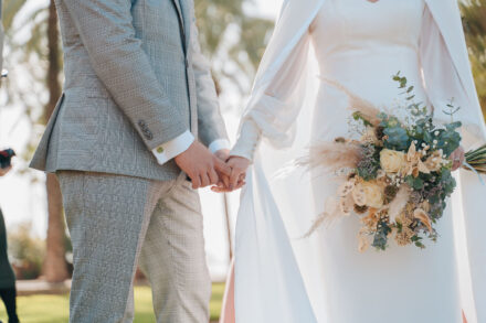 Close-up of the couple holding hands, him in a grey check suit and her holding the dried-flower bouquet