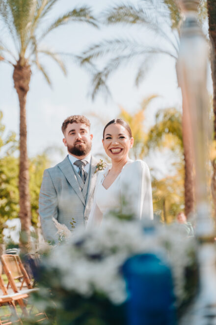 The bride smiles at the camera beside the groom during the ceremony, behind a blurred arrangement of white flowers