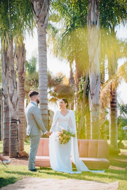 The couple hold hands in front of the pink sofa beneath palm trees, with sunlight streaming through the fronds