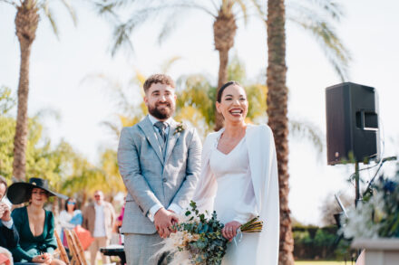 The couple laugh as they stand during the ceremony, his hands clasped and her bouquet in hand, before the guests
