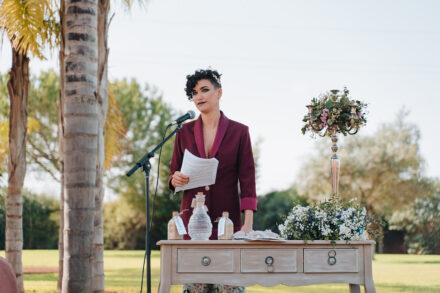 The officiant, in a burgundy blazer, reads at the microphone behind a table with bottles of sand and white flowers