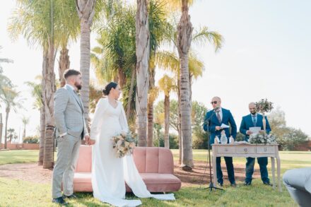 Two guests in blue suits read at the microphone while the couple listen, standing by the pink sofa