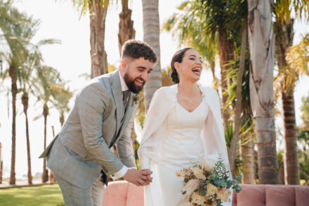 The bride bursts out laughing and the groom laughs leaning towards her, hand in hand in front of the pink sofa