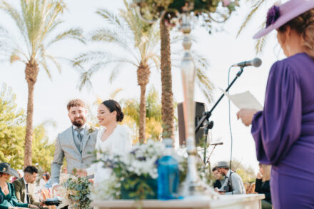 A guest in purple with a wide-brimmed hat reads at the microphone as the smiling couple listen in the background