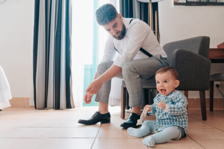 The groom, seated in braces, plays with the baby, who laughs on the floor with a toy giraffe